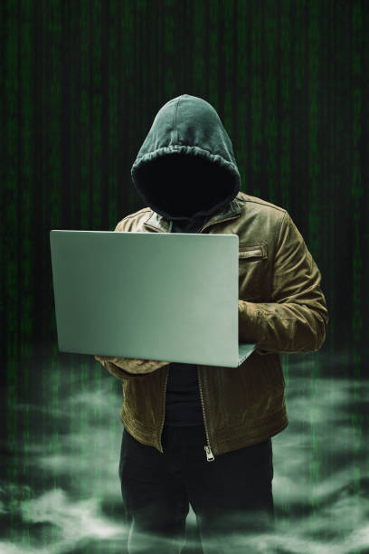 istock Faceless man, hooded cybercriminal terrorist, anonymous hacker standing and holding laptop on a dark background with a smoke 2167112618