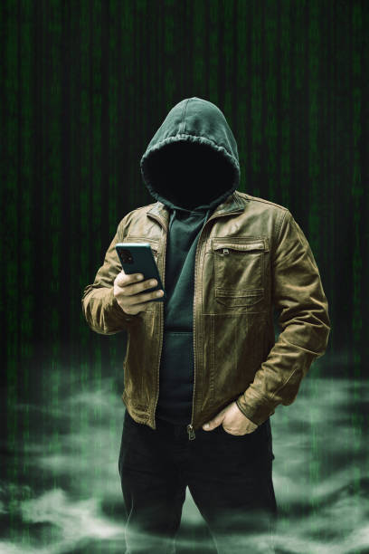 istock Faceless man, hooded cybercriminal terrorist, anonymous hacker standing and using smartphone on a dark green background with a smoke 2167112626