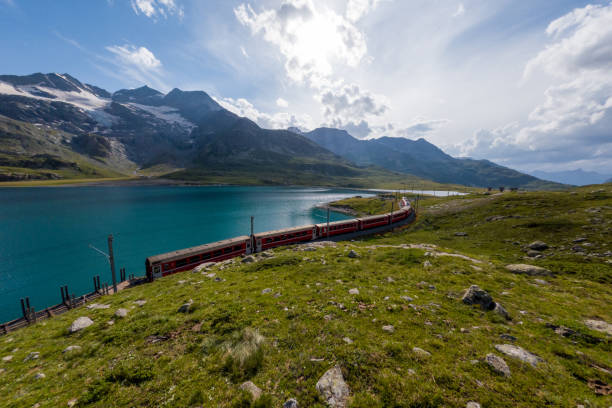 istock Red Train Curving Alongside Scenic Alpine Lake with Mountain Backdrop 2167128450