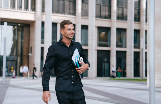 istock young man in a black shirt walks confidently while holding documents and a smartphone, embodying a successful and focused professional on the move in an urban setting 2167129385