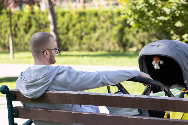 Father with a stroller in a park sitting on a bench and relaxing until baby is sleeping. Rear view
