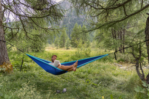 A man enjoys a leisurely moment in a hammock by a stream, reading a book and surrounded by the tranquility and lush greenery of a forest.