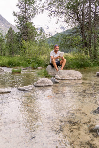 istock Man Reading a Book While Sitting on a Rock in a Mountain Stream 2167132672