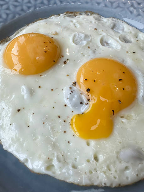 istock Full frame image of blue plate with floral patterned rim containing two merged cooked sunny side up fried eggs seasoned with cracked pepper corn, broken oozing yolk. elevated view, focus on foreground 2167141772