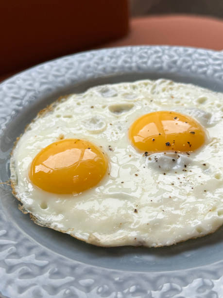 istock Close-up image of blue plate with floral patterned rim containing two merged cooked sunny side up fried eggs seasoned with cracked pepper corn, elevated view, focus on foreground 2167141781