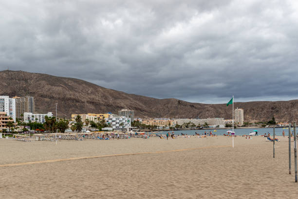 istock Cloudy day at a tranquil beach with a mountain backdrop, empty sun loungers, and a green flag indicating safe swimming conditions in Los Cristianos, Tenerife. 2167150075
