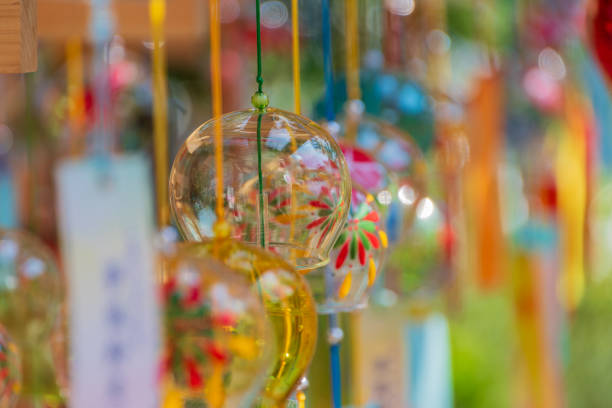 A mesmerizing close-up of traditional Japanese glass wind chimes, adorned with intricate floral patterns. The colorful chimes, hung in rows, reflect the soft light, creating a warm and inviting glow. Each chime is attached to a paper strip that gently sways in the breeze, filling the air with the soothing, melodic sounds unique to summer festivals in Japan. The bokeh effect in the background adds a dreamy atmosphere, enhancing the overall sense of peace and nostalgia.