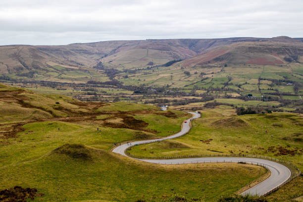 istock Winding road through a lush, green valley with rolling hills under a cloudy sky in Peak District, England. 2167171106