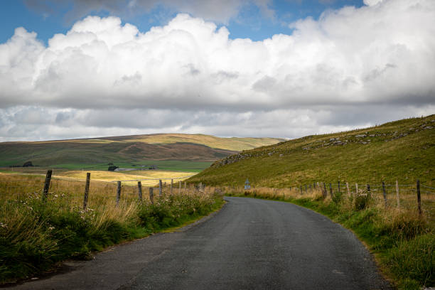 istock Rural road leading through rolling hills under a cloudy sky, with green fields and wooden fence on the side in Yorkshire Dales, England. 2167184175