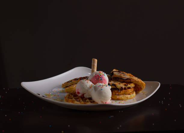 Waffles served with strawberry and vanilla ice cream, colorful sprinkles, a waffle cone, and chocolate sauce, on a white plate with a dark background.