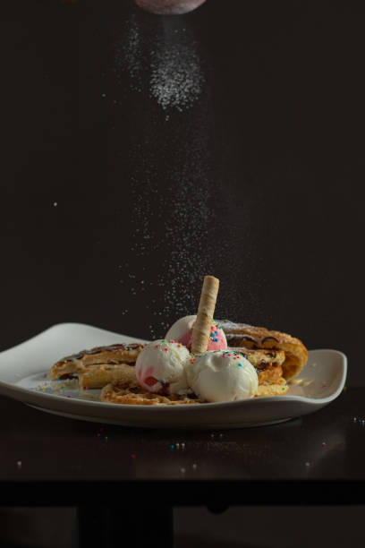 Waffles with strawberry and vanilla ice cream, colorful sprinkles, waffle cone, and chocolate sauce, with powdered sugar falling from above, served on a white plate with a dark background.