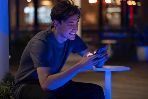 istock Cheerful young man using phone sitting on bench on street at night 2167193594