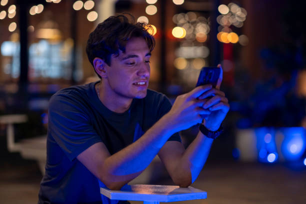 istock Cheerful young man using phone sitting on bench on street at night 2167193606