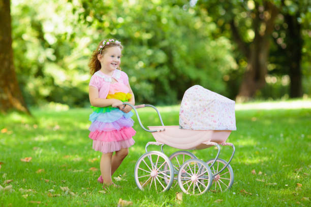 Little girl pushing toy stroller with bear. Toddler kid in pink dress playing with doll buggy. Kids birthday party. Children play outdoors. Mother and baby role game. Family summer fun. Preschool toys