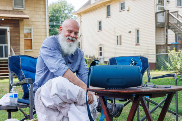 Senior beekeeper with a long white beard relaxes outdoors after completing hive inspection. He sits in a blue camping chair, protective gear removed, dressed in a light blue shirt and casual pants. Residential buildings in the background indicate a backyard or community apiary setting.