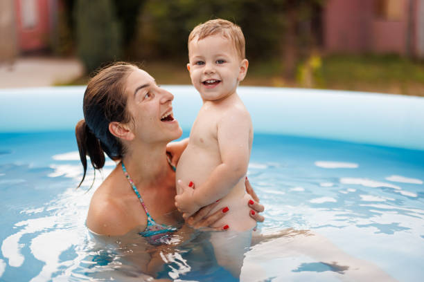 Smiling baby boy looking at camera while mother hugging him baby in wading pool on sunny day