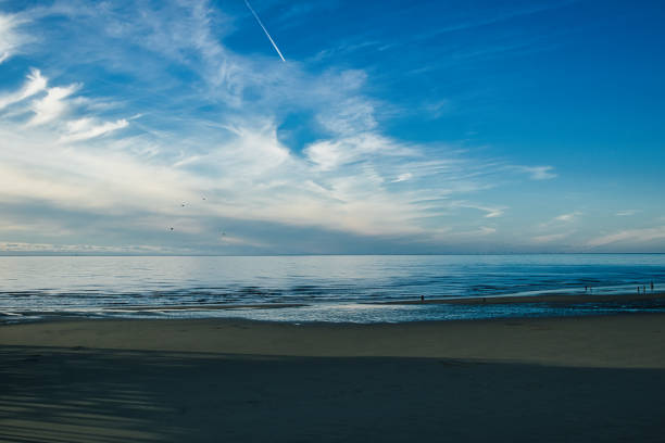 Serene beachscape with calm blue waters under a vast sky with wispy clouds and a distant airplane trail.