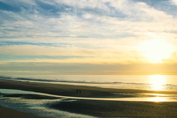 istock Serene beach sunrise with reflective tidal pools and a distant figure walking along the shore. 2167260027