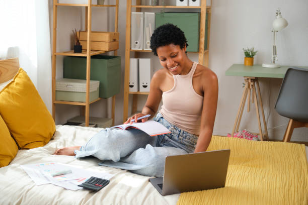 istock Young black woman working from home sitting on bed using laptop and taking notes 2167273792