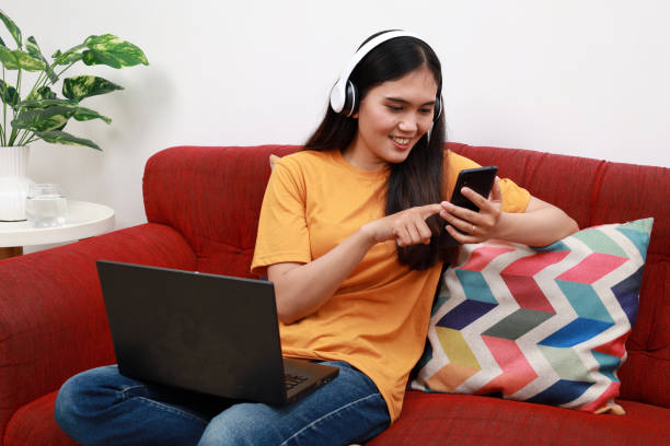 istock Young Asian woman sitting on sofa at home using mobile phone to send text messages and talk on the phone with her friends. 2167274824