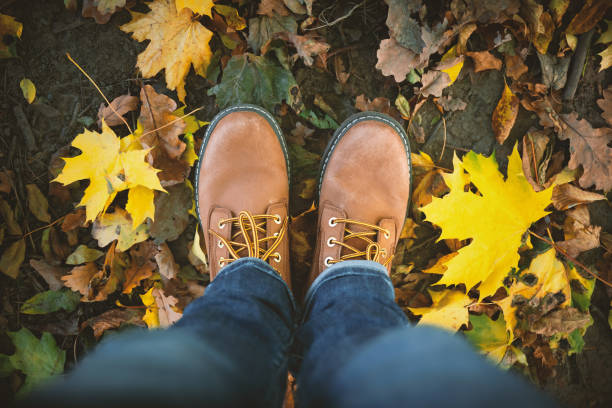 Man legs in jeans and brown leather boots on park ground with colorful fallen maple leaves around. Concept of fall autumn season and trendy hipster lifestyle