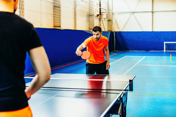 A focused man in an orange shirt is preparing to hit the ball in an indoor table tennis match, with his opponent visible in the foreground