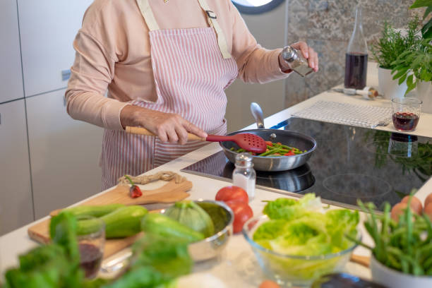 istock Elderly woman at home cooking vegetables in a pan while adding some pepper. Elderly lady in the kitchen preparing dinner. Healthy eating concept 2167286956