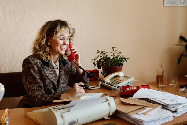 Professional woman organizing office documents while holding a retro phone, against a blue background in a casual yet stylish work environment