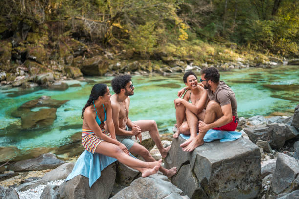 Cheerful young multiracial couples delighting in a leisurely respite amid the beauty of nature. Perched on stones beside a river adorned with turquoise waters, they opt for swimsuits to combat the summer warmth.