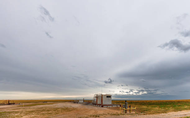 Lone oil well stands in vast, open prairie landscape of Canadian County, Oklahoma. White structure and metal equipment contrast against golden grasslands stretching to horizon. Dramatic sky filled with gray clouds creates moody atmosphere, capturing essence of Oklahoma's energy industry amidst natural beauty.