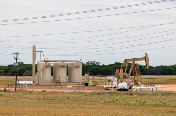 Extracting oil in Blaine County. Pump jack and storage tanks dominate rural Oklahoma grassland. Pumpjack actively extracts oil alongside large cylindrical storage tanks in rural Blaine County, Oklahoma. Power lines stretch across the sky above industrial equipment. Oil field situated on flat grassland with distant trees, typical of Oklahoma landscape. oil wellhead stock pictures, royalty-free photos & images