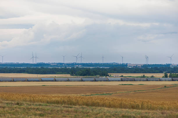 Panoramic rural landscape showcases renewable energy sources alongside traditional farming. Numerous wind turbines dot the distant horizon against a cloudy sky with freight train passing by. Middle ground features a row of solar panels reflecting daylight. Foreground displays golden wheat or barley fields ready for harvest, illustrating the integration of sustainable energy production with agricultural practices.