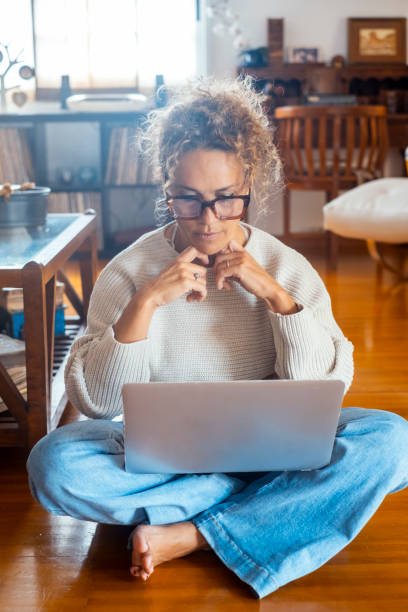 istock Modern woman in smart working thinking for a solution sitting on the floor crossed legs with laptop looking at the screen. Remote worker in living room wearing casual clothes and glasses. Online job 2167341375