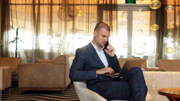istock Focused businessman working on a laptop computer in a lounge interior. A man talks on a smartphone at a remote workplace. Freelancing and online learning concept. 2167369714