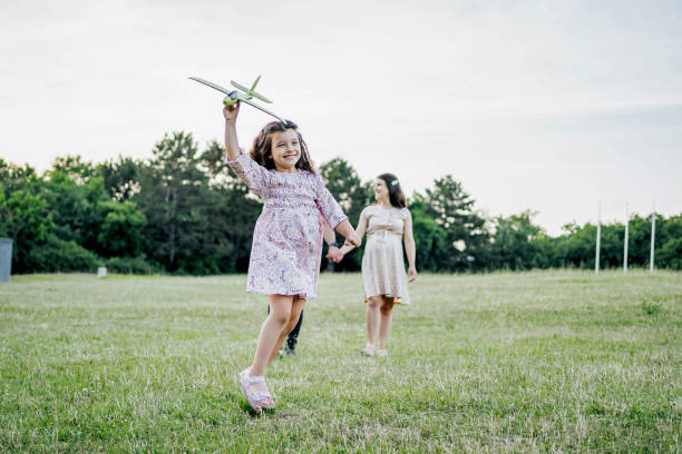 Cute little girl running through the meadow on a sunny day with a toy plane in hand. Happy kid playing with cardboard plane against blue summer sky background. Childhood dream imagination concept.