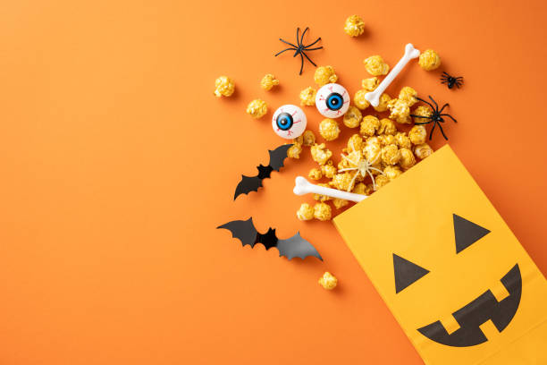 istock Halloween-themed popcorn with spooky decorations like bats, eyes, and bones spilling from a pumpkin bag on an orange background 2167406177