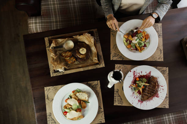 istock Set of dishes from: Caesar salad, grilled eggplant steak, vegetable salad with poached egg and Striploin steak in a wine sauce. Top view of woman having sitting at the wooden table 2167412402