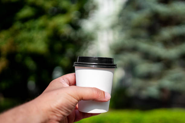 A close-up of a hand holding a white takeaway coffee cup with a black lid, with a blurred tree in the background on a sunny day. Perfect for themes of coffee, outdoor lifestyle, and relaxation.