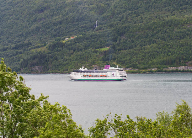 A ferry cruises along the tranquil waters of Innvikfjorden, surrounded by lush, green mountains in Stryn, Norway. Snow-capped peaks tower above, creating a picturesque and serene landscape.