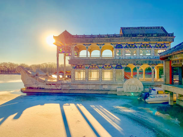 The Marble Boat, also known as the Boat of Purity and Ease, is illuminated by the warm, golden hues of the setting sun at the Summer Palace, Beijing, China. This iconic Qing Dynasty structure, made of marble and wood, stands majestically over the frozen lake, with long shadows and tranquil reflections capturing the serene winter evening.