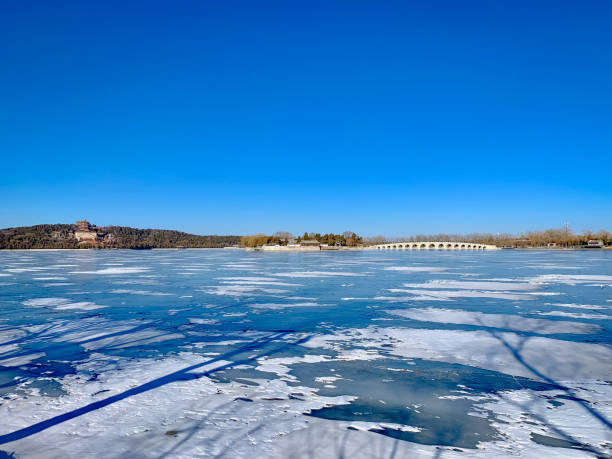 A breathtaking winter scene featuring Longevity Hill and the Seventeen-Arch Bridge across a frozen Kunming Lake at the Summer Palace in Beijing, China. The bright blue sky and icy surface of the lake contrast with the historic architecture, creating a serene and timeless view of this UNESCO World Heritage Site.
