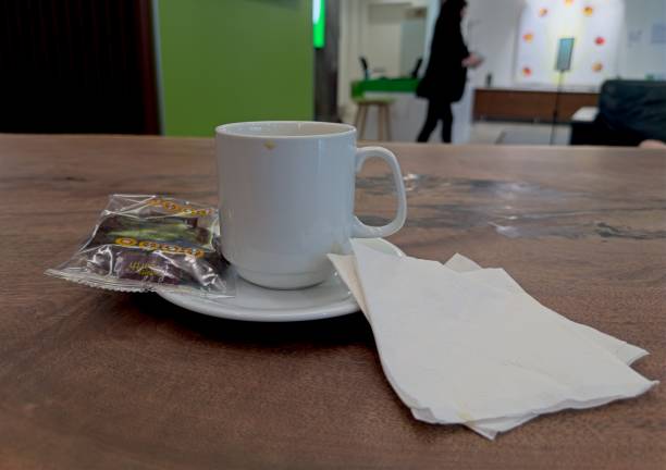 A white coffee cup on a saucer with a small packet of biscuits and napkins on a wooden table. In the background, a blurred figure is walking in a modern indoor space with green and white decor.