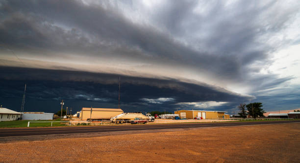 Dramatic supercell thunderstorm approaches a small Oklahoma town, featuring a massive, dark shelf cloud dominating the sky. Storm's leading edge forms a distinct horizontal line, contrasting with lighter sky beyond. Industrial buildings and structures stand quietly under the impending storm, highlighting vulnerability against nature's raw power.
