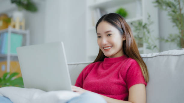 istock Happy young Asian woman using laptop while seated on couch at home 2167488392