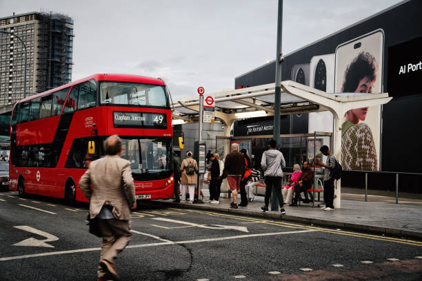 Ariel Way / Shepherd's Bush Bus Station, west London, England, United Kingdom – 08/19/2024. pedestrian traffic from commuters and shoppers: People, Shoppers, & Commuters waiting at bus stops and navigating the area. Shepherd's Bush train station building (Shepherd's Bush Overground Station; a sleek glass and metal structure) in the background at Ariel Way, near Shepherd's Bush station, red double-decker bus on the street.