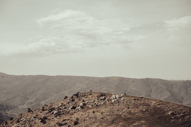 A mountain range with a cloudy sky in the background. The sky is mostly gray with a few white clouds scattered throughout. The mountains are covered in rocks and dirt, giving the scene a rugged