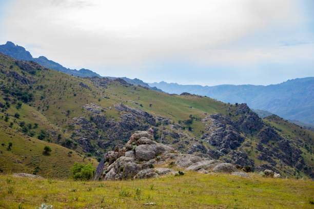 A mountain range with a rocky hillside and a grassy field. The sky is cloudy and the mountains are in the background