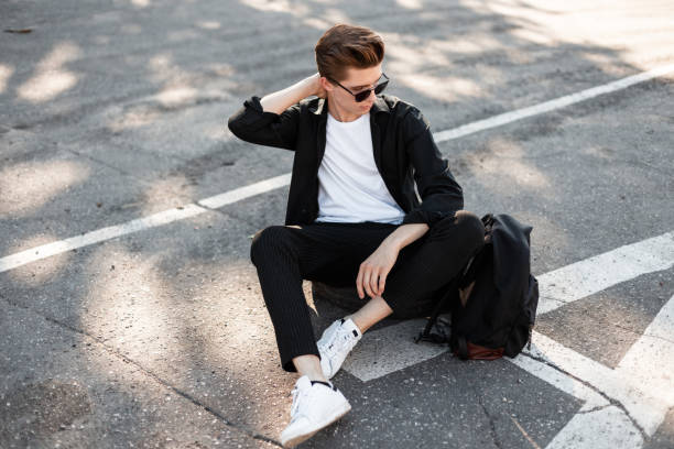 istock Nice young man hipster in elegant black clothes in white trendy sneakers with a backpack in sunglasses sits on a rubber wheel on the street. Cool guy relaxes in the sun outdoors on a summer day. 2167519232