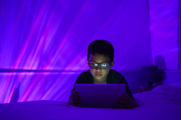 istock A young boy wearing glasses is using a tablet while sitting on a bed in a room with purple and blue ambient lighting. 2167527994