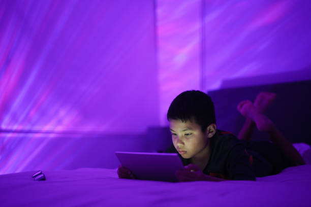 Boy lying on bed using a tablet with a glowing purple light in a dark room creating a magical ambiance.
